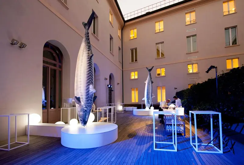 Pesci giganti e tavolini luminosi in un cortile di un palazzo, di sera